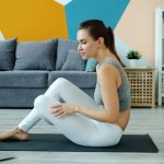 Young lady in top and leggings is using laptop to learn yoga asanas watching tutorial exercising at home on mat. Modern technology and millennials concept.