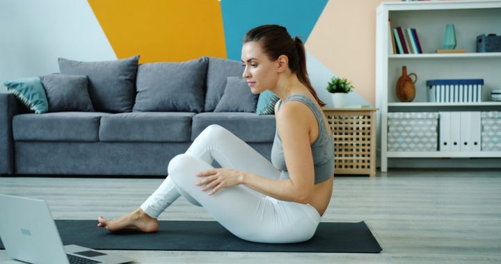 Young lady in top and leggings is using laptop to learn yoga asanas watching tutorial exercising at home on mat. Modern technology and millennials concept.